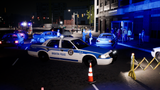 A nighttime street scene with a white and blue Brighton Police car in the foreground, blue emergency lights on, and several people standing near a building entrance in the background under artificial blue lighting.